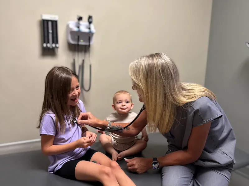 A Health First provider listening to a school-age girl’s chest with a stethoscope during a check-up, her baby sister sitting beside her on the exam table